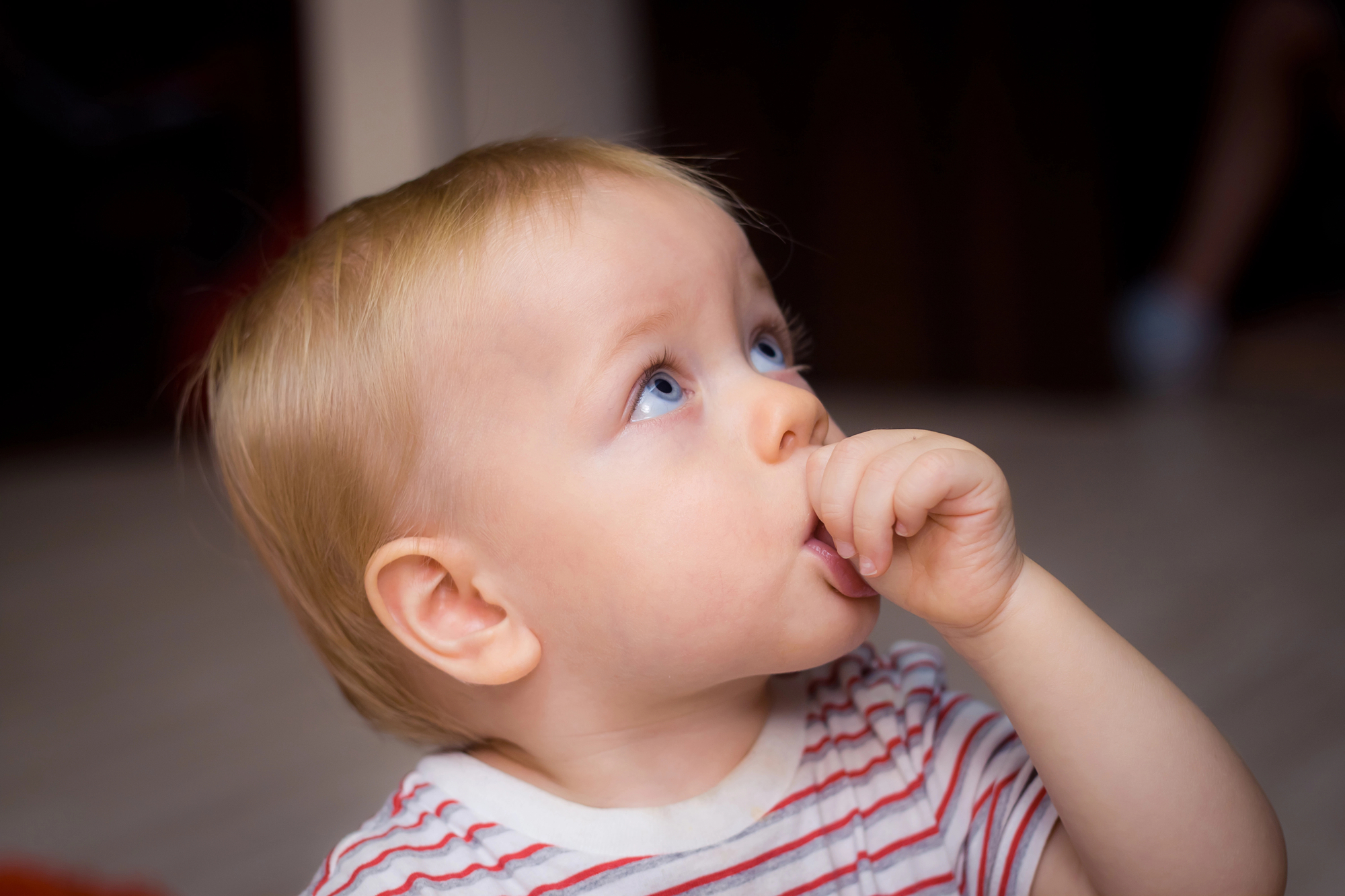 Toddler with wide eyes sucking thumb indoors.