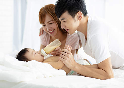 happy father and mother feeding baby from bottle A man and woman feeding a baby on a bed.