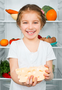 A little girl holding a piece of cheese in front of an open refrigerator.