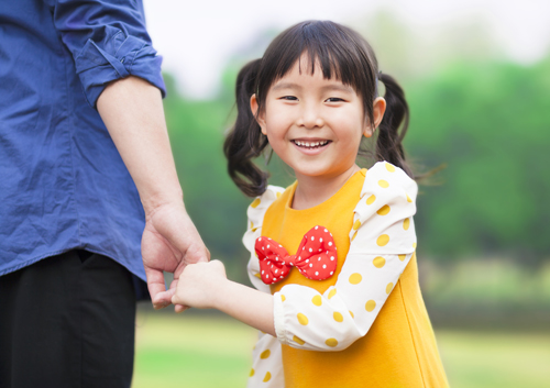 A young girl is holding the hand of her father in a park.
