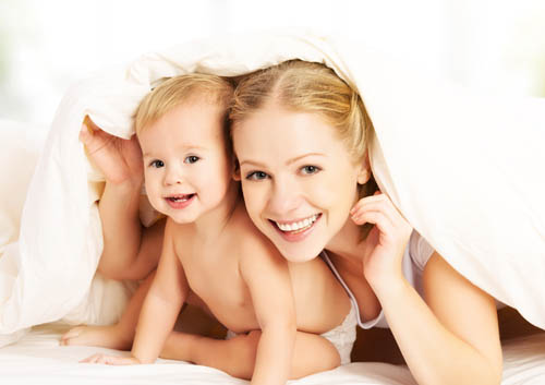 A mother and baby laying under a blanket in bed.