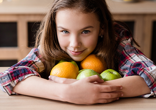 A girl is laying on a table with a bunch of oranges and apples.